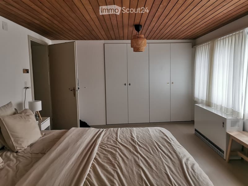 Modern bedroom with a large bed, nightstand, lamp, and built-in white wardrobe. Wooden ceiling and large window with white curtains.