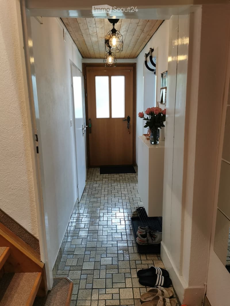 A narrow hallway with tiled flooring, white walls, and a wooden door at the end. A small console table with a vase of flowers is visible on the right side, along with a shoe rack holding several pairs of shoes and sandals.