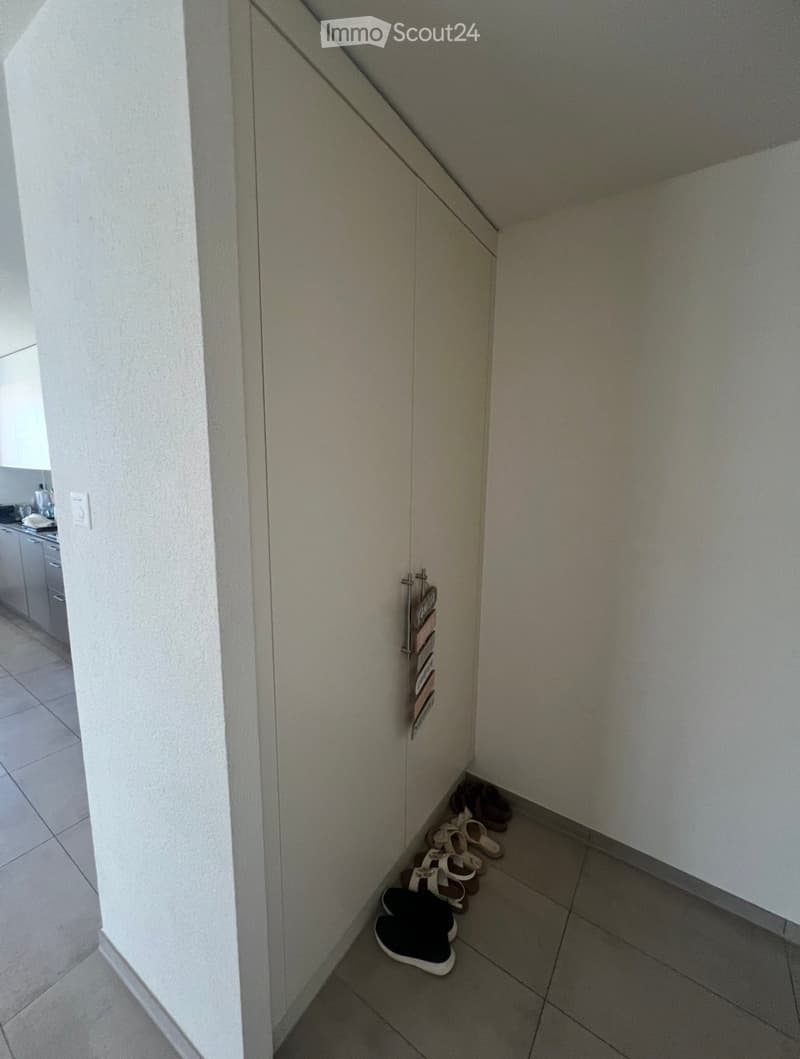 A narrow hallway with tiled flooring, white walls, and a small shoe rack holding several pairs of shoes. The hallway leads to a kitchen area visible in the background.