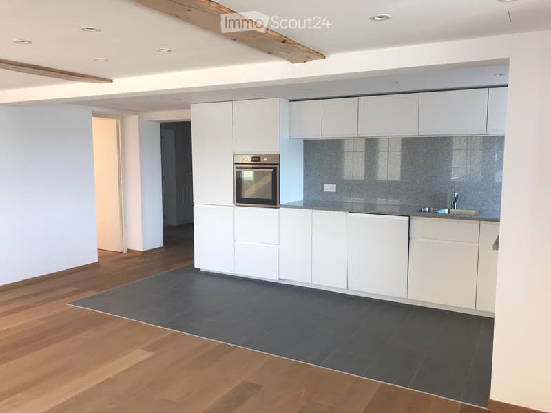 Modern kitchen with white cabinets, dark grey tiles, wooden flooring, and an integrated oven. The space is clean and well-lit with exposed wooden beams on the ceiling.