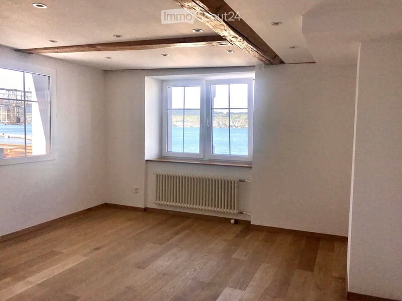 Empty room with wooden flooring, white walls, exposed wooden beams on the ceiling, and two windows offering a view of the sea.