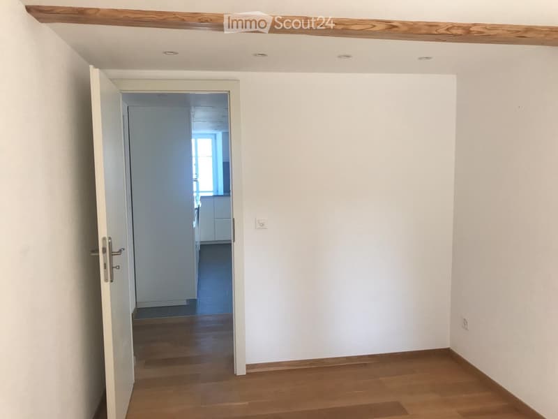 Empty room with wooden flooring, white walls, and an open door leading to another room with white cabinets and a window.