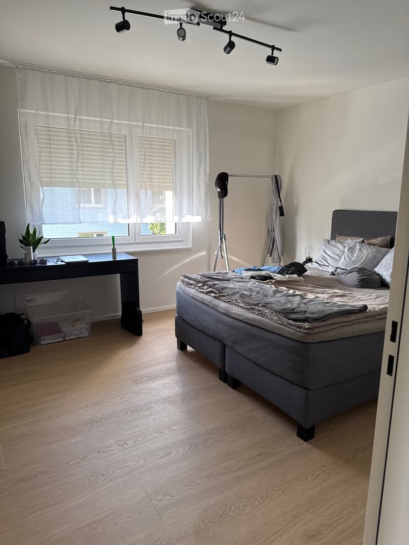 Modern bedroom with a grey bed, wooden floor, large window with blinds, desk with laptop and plant, and a mirror on a stand.
