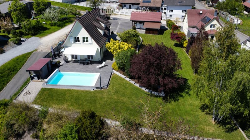 Aerial view of a modern house with a swimming pool, surrounded by a well-maintained lawn and trees. The house features a white exterior with a dark roof and a covered patio area with outdoor furniture.