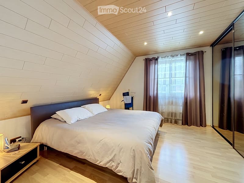 Modern bedroom with a large bed, nightstand, lamp, and large window with curtains. The room features wooden flooring and a slanted ceiling with built-in lighting.