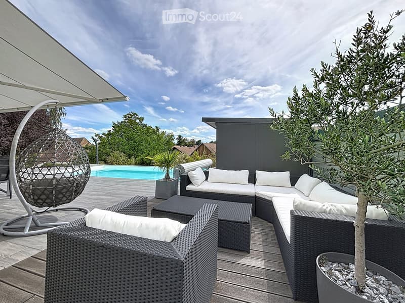 A modern outdoor patio with a wicker sofa, chairs, a hanging egg chair, a small table, and a potted plant, overlooking a swimming pool and garden area.