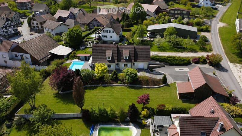 Aerial view of a residential neighborhood with multiple houses, well-maintained lawns, trees, and two swimming pools visible in the foreground.