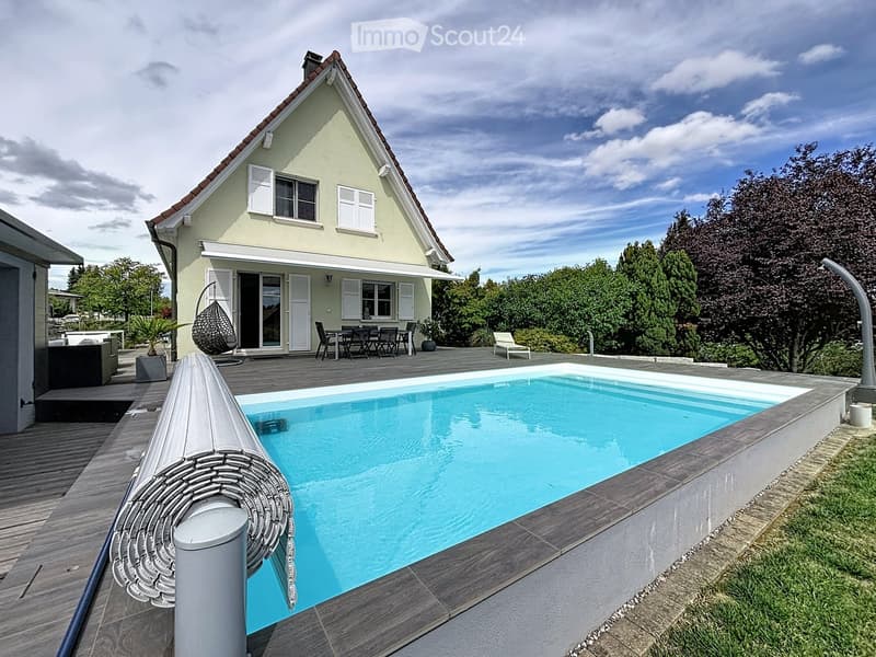 Outdoor area with a swimming pool, wooden deck, outdoor furniture, and a modern house with white shutters and a sloped roof.