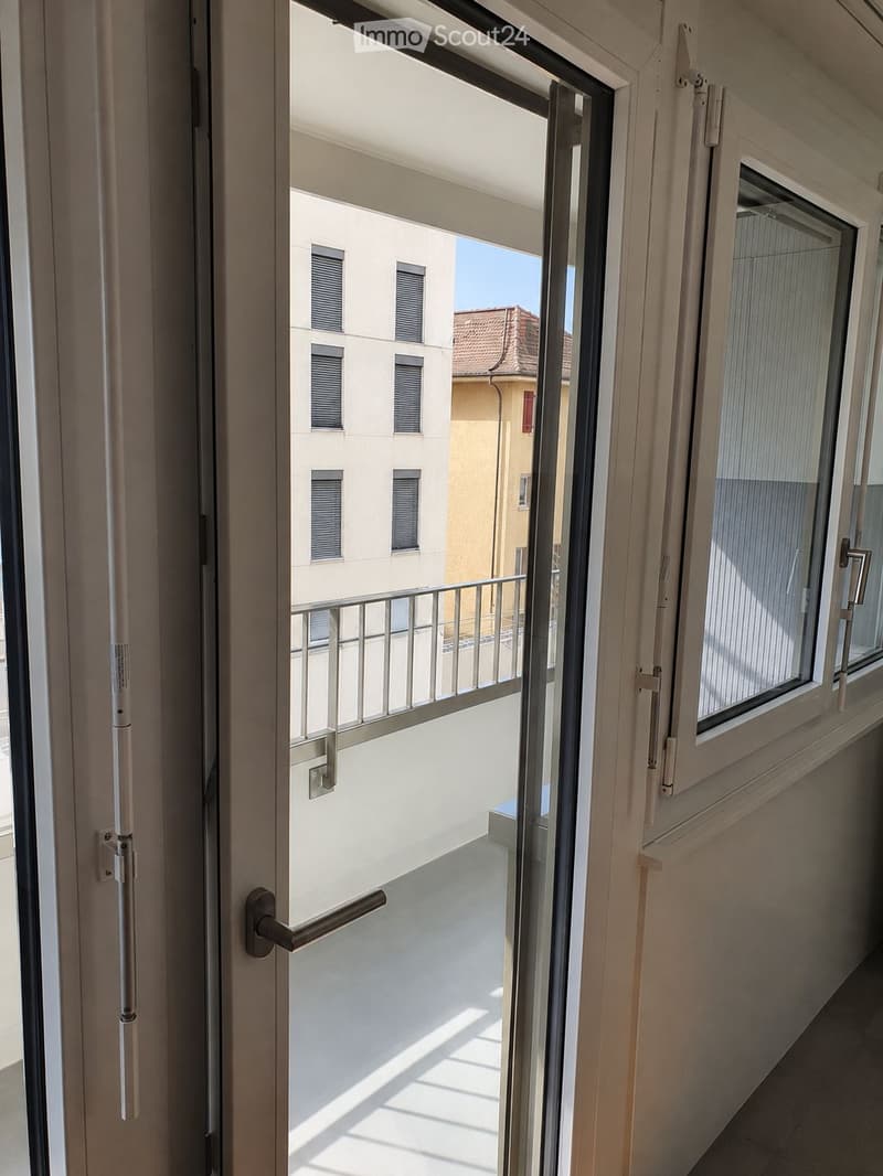 Open sliding door leading to a small balcony with a metal railing, overlooking neighboring buildings.