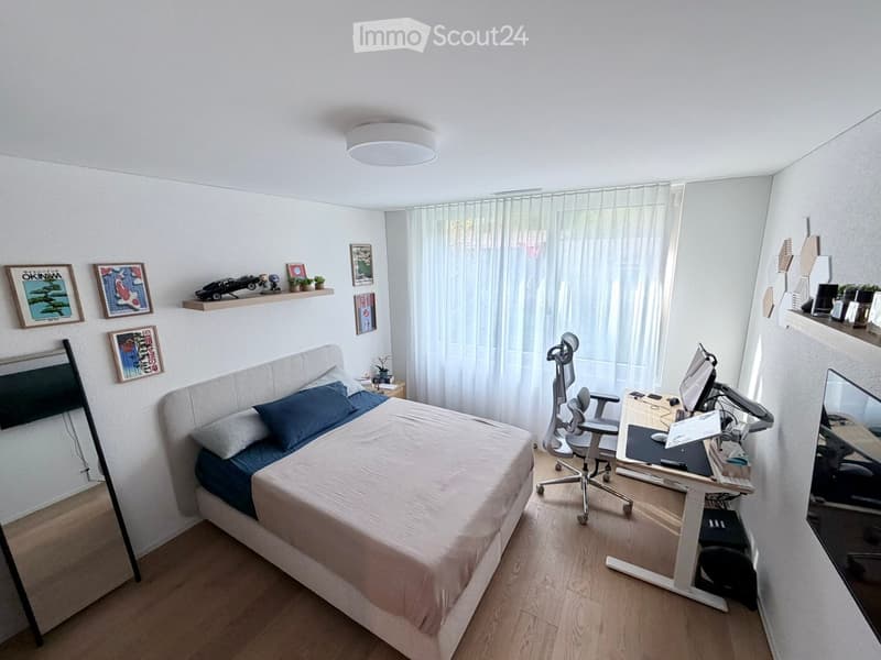 A modern, well-lit bedroom with a bed, desk, and shelves. The room features a large window with white curtains, a sleek desk setup with a computer, and decorative shelves with framed pictures and small plants.