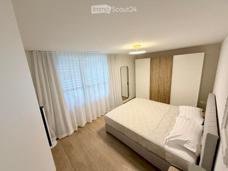Cozy bedroom with a single bed, white bedding, a wardrobe, and a mirror. Light wooden flooring and large windows with white curtains.