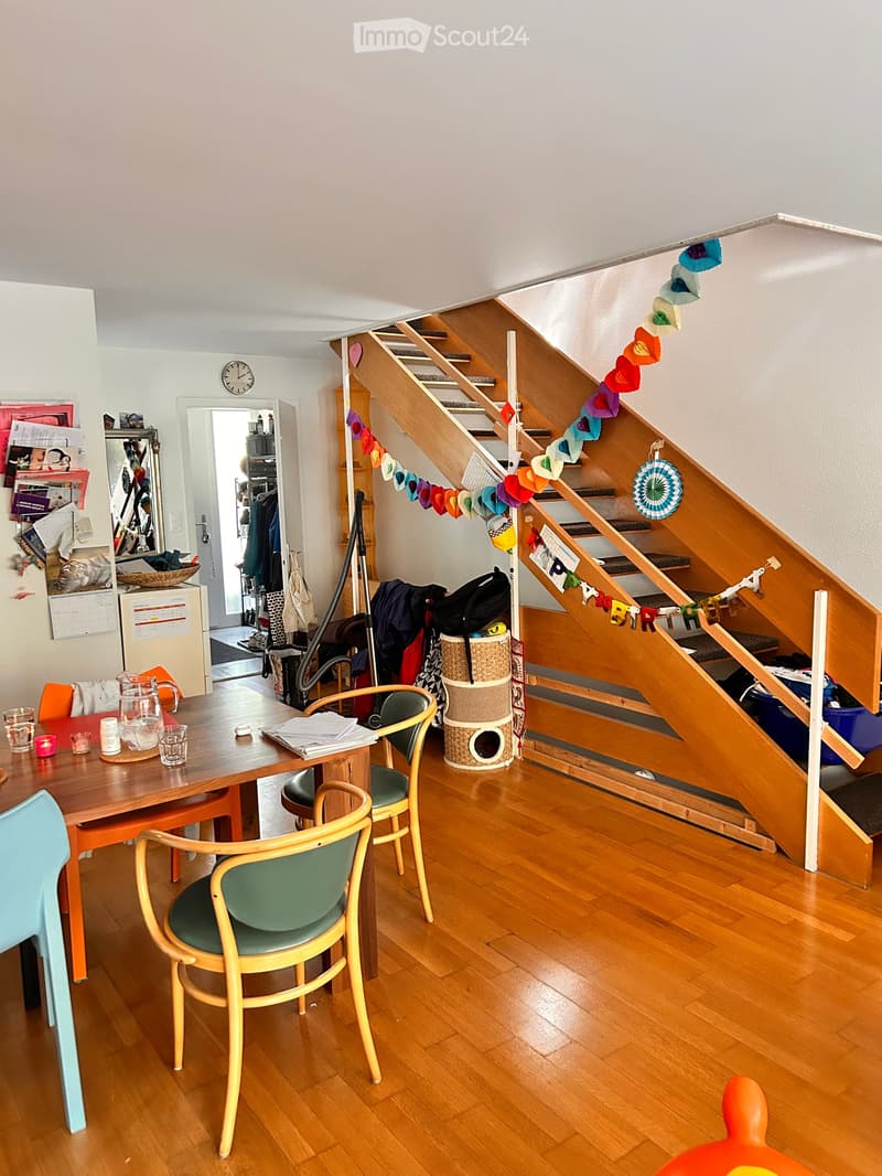 A cozy living space with a wooden staircase, a dining table with chairs, a cat tree, and a refrigerator in the background.
