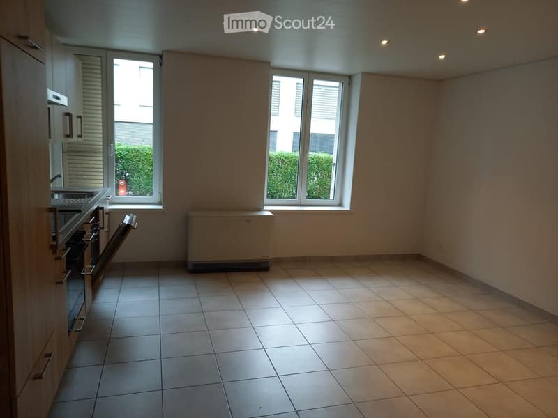 Empty kitchen with tiled floor, white walls, and large windows. Features a sink, oven, and radiator.