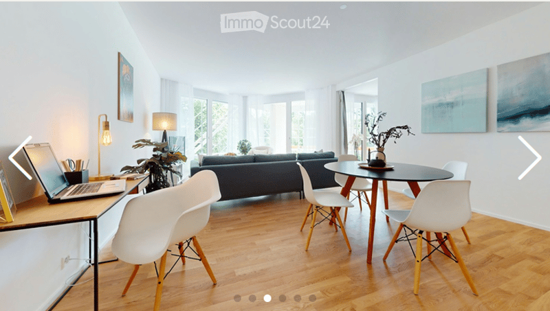 Modern living room with a dark blue sofa, white chairs, a round dining table, a laptop on a desk, and decorative plants and artwork on the walls.