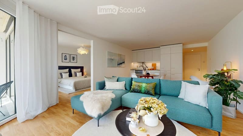 Modern, open-concept living and bedroom area with a blue sectional sofa, coffee table, bed with pillows, kitchen area with cabinets, and decorative items.