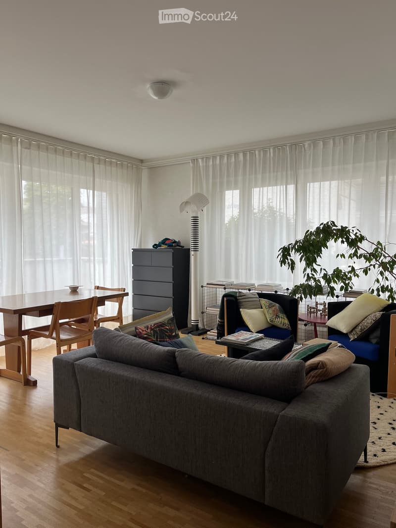Bright living room with large windows covered by sheer curtains, wooden flooring, grey sofa with colorful pillows, coffee table, dining table with chairs, floor lamp, and a tall plant in the corner.