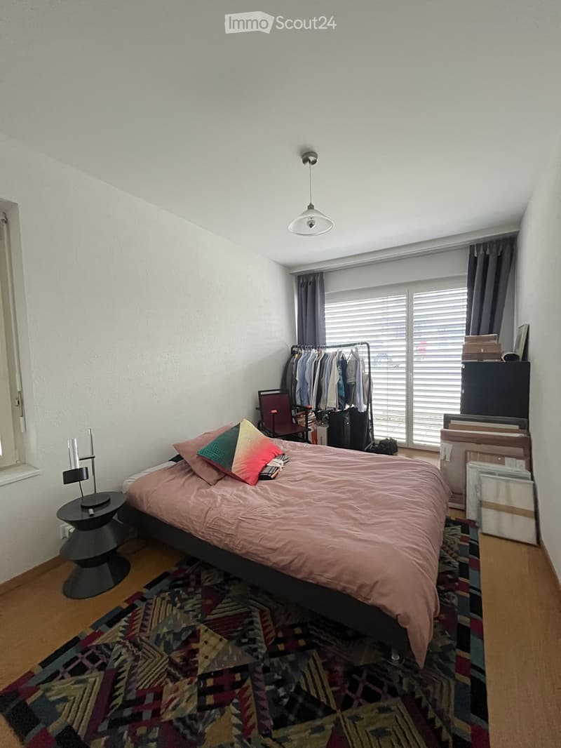 A modern bedroom with a double bed, colorful pillows, a nightstand with a lamp, a chair, a clothing rack, and a suitcase near the window.