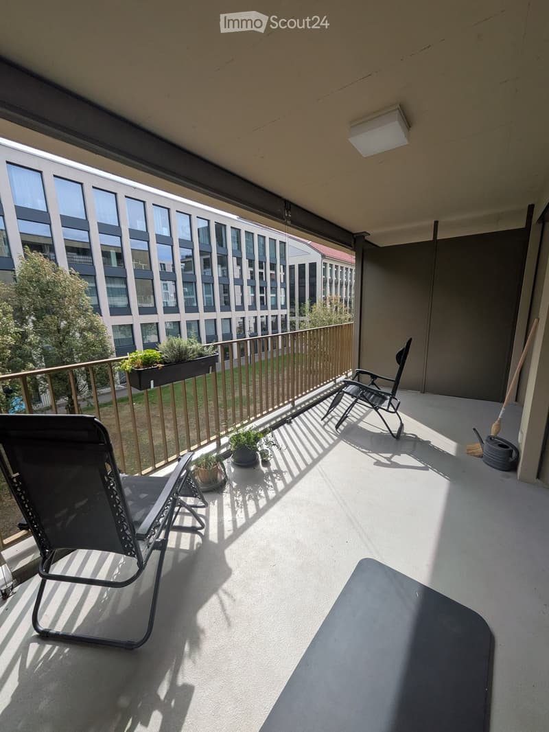 Balcony with two black folding chairs, small table, potted plants, and a railing overlooking a modern building.