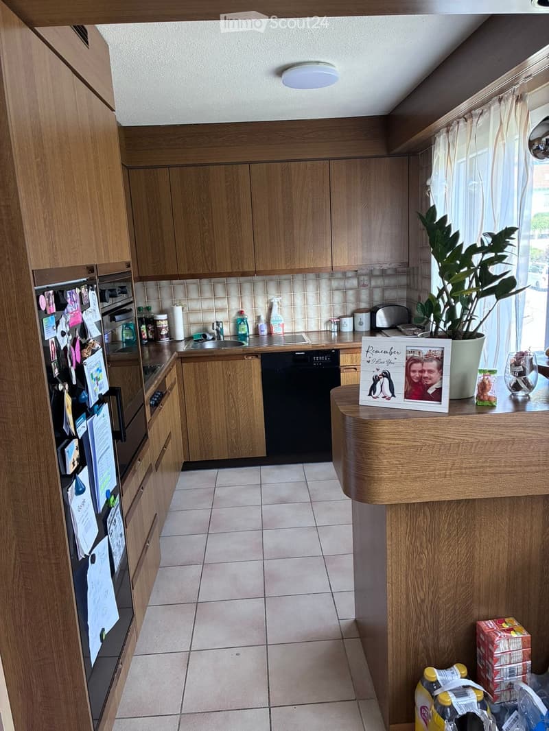 Well-lit kitchen with wooden cabinets, stainless steel appliances, and a central island. Visible items include a refrigerator, oven, dishwasher, sink, toaster, and a framed photo on the counter.