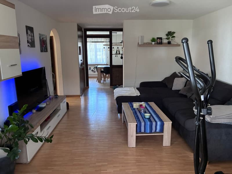 Modern living room with wooden flooring, a dark grey sectional sofa, a light wooden coffee table with a blue and white runner, a TV stand with a flat-screen TV, and a potted plant. An exercise bike is positioned near the sofa.