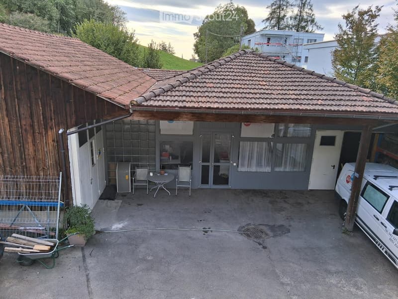 Outdoor area with a small covered patio, a table and chairs, a van parked nearby, and a wheelbarrow on the left side.