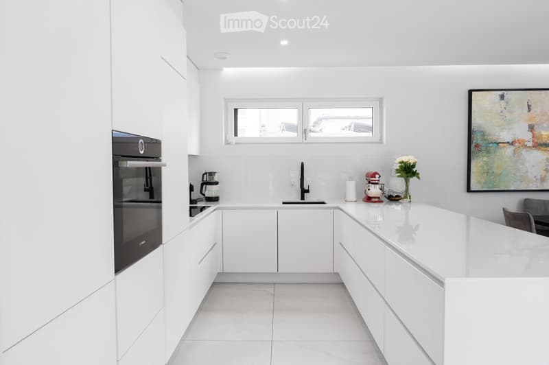 Modern white kitchen with sleek cabinetry, a built-in oven, a central island, and a vase with flowers on the counter.
