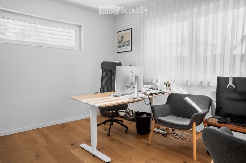 Modern, well-lit room with a standing desk, ergonomic chair, laptop, keyboard, headphones, and a comfortable seating area with a TV and additional chair.
