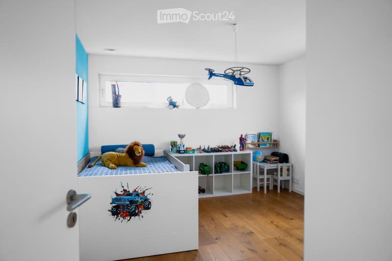 A child's bedroom with a bed, a large stuffed lion toy, shelves filled with books and toys, a small table and chairs, and a globe. The room features wooden flooring and white walls with a blue accent wall.