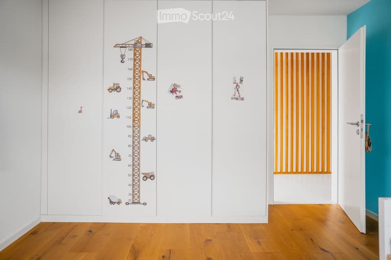 A narrow hallway with white walls, wooden flooring, and a height chart mural featuring construction vehicles. A door leads to another room with orange vertical blinds.