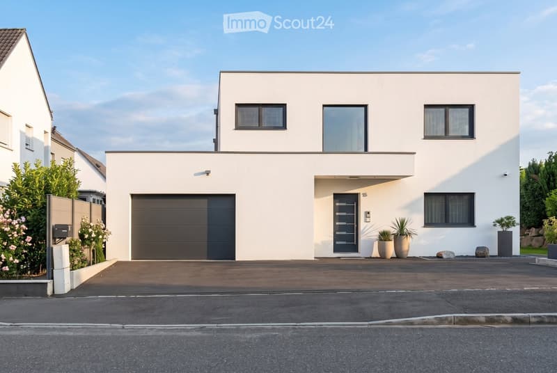 Modern two-story house with white exterior, large garage, and contemporary design. Features include large windows, a raised ground floor, and a well-maintained driveway.