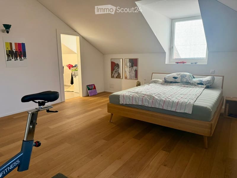 Cozy attic bedroom with wooden flooring, a single bed with a patterned quilt, a nightstand, a bicycle, and several picture frames on the walls.