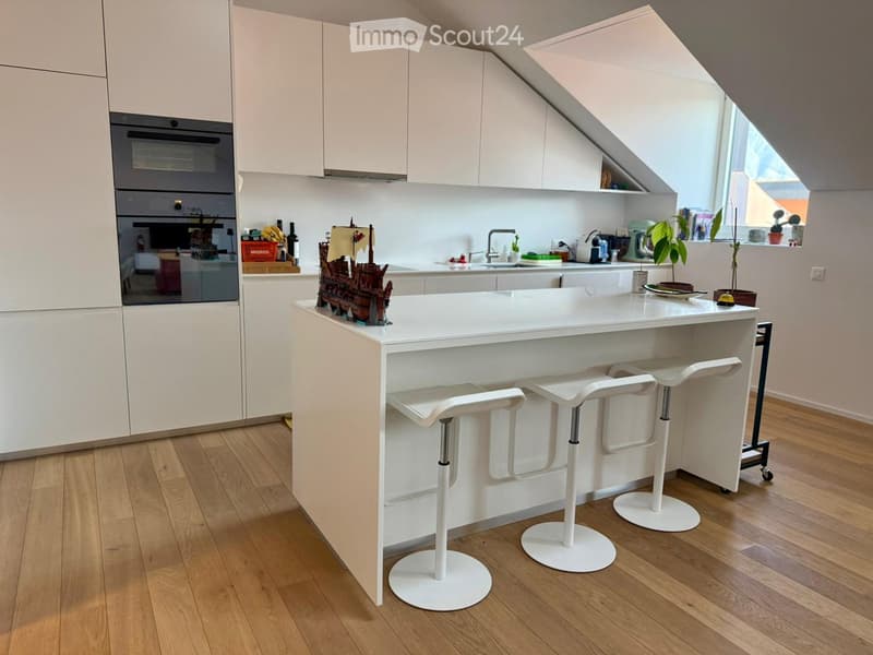 Modern kitchen with white cabinets, central island, and four white bar stools. Features an oven, sink, and various kitchen items on the counter.