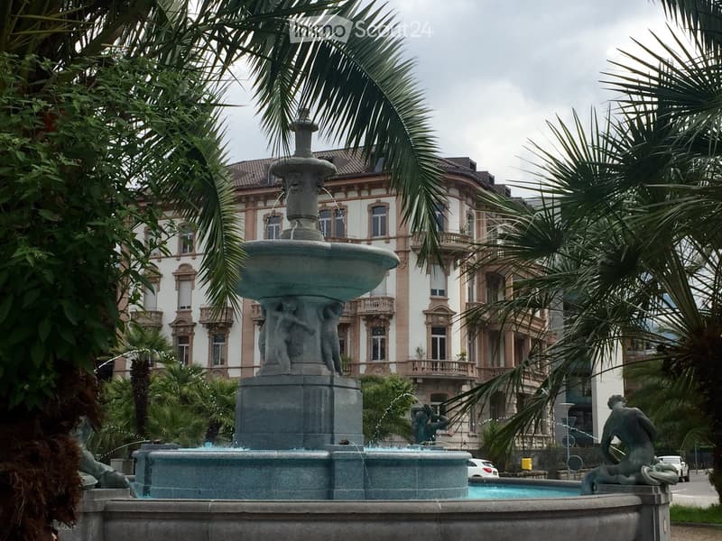 A large stone fountain with multiple tiers and sculptures, surrounded by palm trees and a grand historic building in the background.