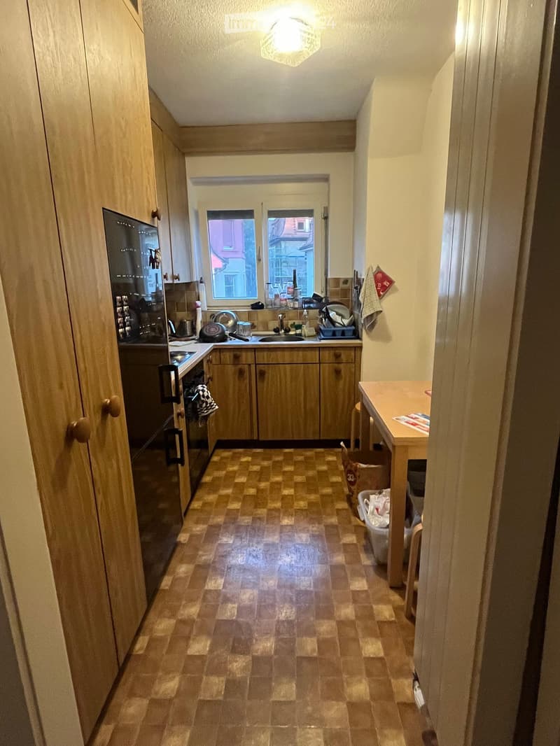 A compact kitchen with wooden cabinets, a tiled floor, and a small table near the window. The kitchen features a sink, oven, and refrigerator, with various kitchen items on the counter.