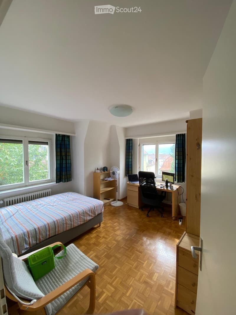 A furnished bedroom with a bed, desk, chair, laptop, fan, and a small chair with a green bag on it. The room has wooden flooring and large windows with curtains.