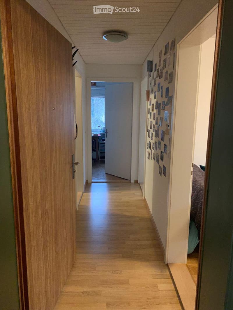 A narrow hallway with wooden flooring and white walls, featuring a collage of photos on one wall and a closed door at the end.