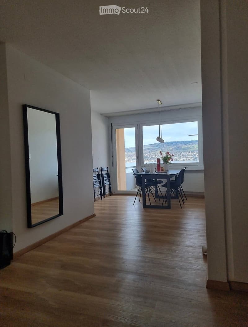 Bright, open dining area with wooden flooring, large windows offering a scenic view, a dining table with chairs, and a decorative flower vase on the table.