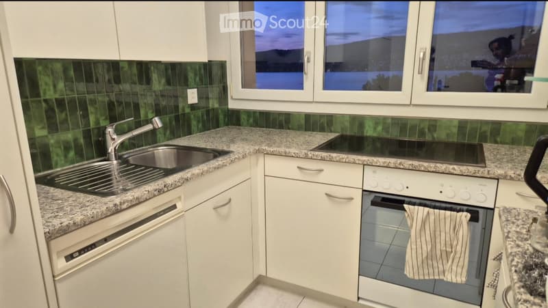 Modern kitchen with white cabinets, granite countertops, stainless steel sink, dishwasher, electric stove, and oven. Green tiled backsplash and large windows.