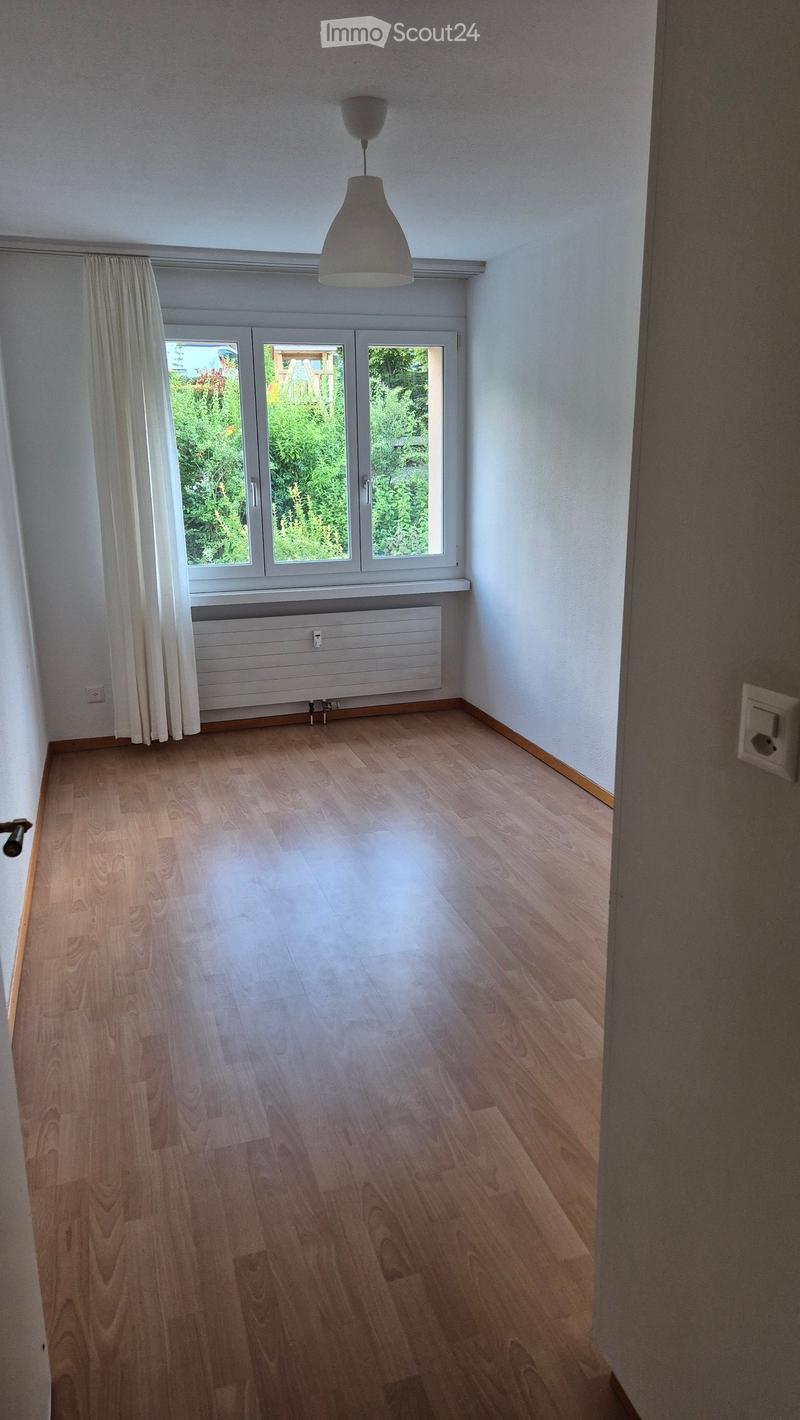 Empty room with wooden flooring, white walls, large windows with white curtains, radiator, and a hanging light fixture.