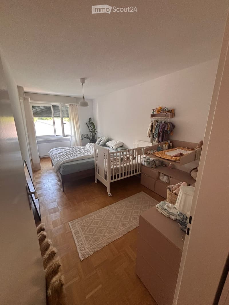Brightly lit bedroom with wooden flooring, a double bed, a crib, a changing table, a wardrobe, and a small plant near the window.