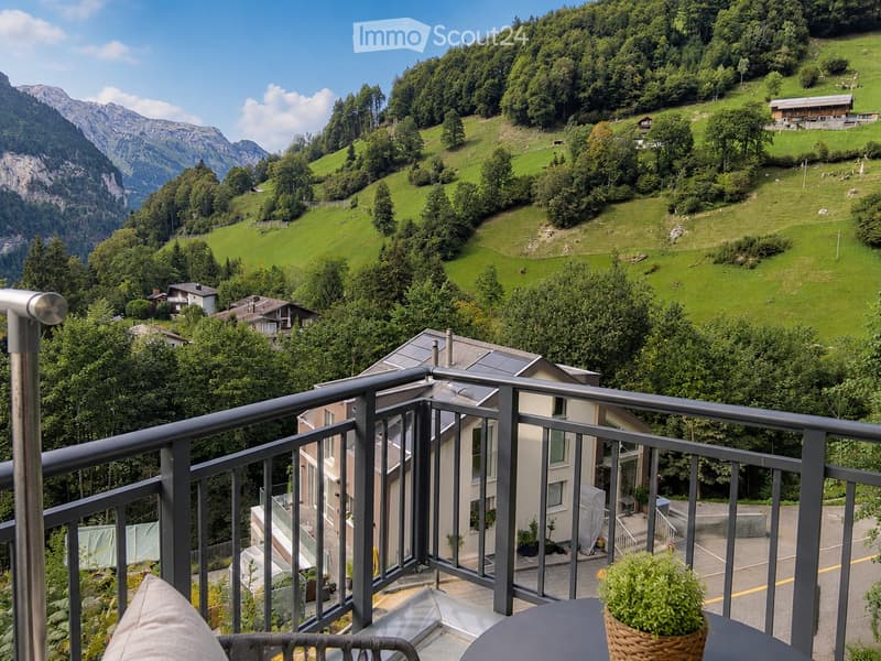 Balcony overlooking a lush green valley with rolling hills, scattered houses, and distant mountains under a clear blue sky.