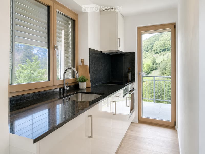 Modern kitchen with white cabinets, black countertops, stainless steel sink, and a view of greenery through large windows and a sliding door leading to a balcony.