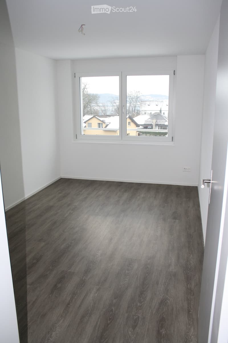Empty room with white walls, dark wooden flooring, and large windows offering a view of the surrounding area.