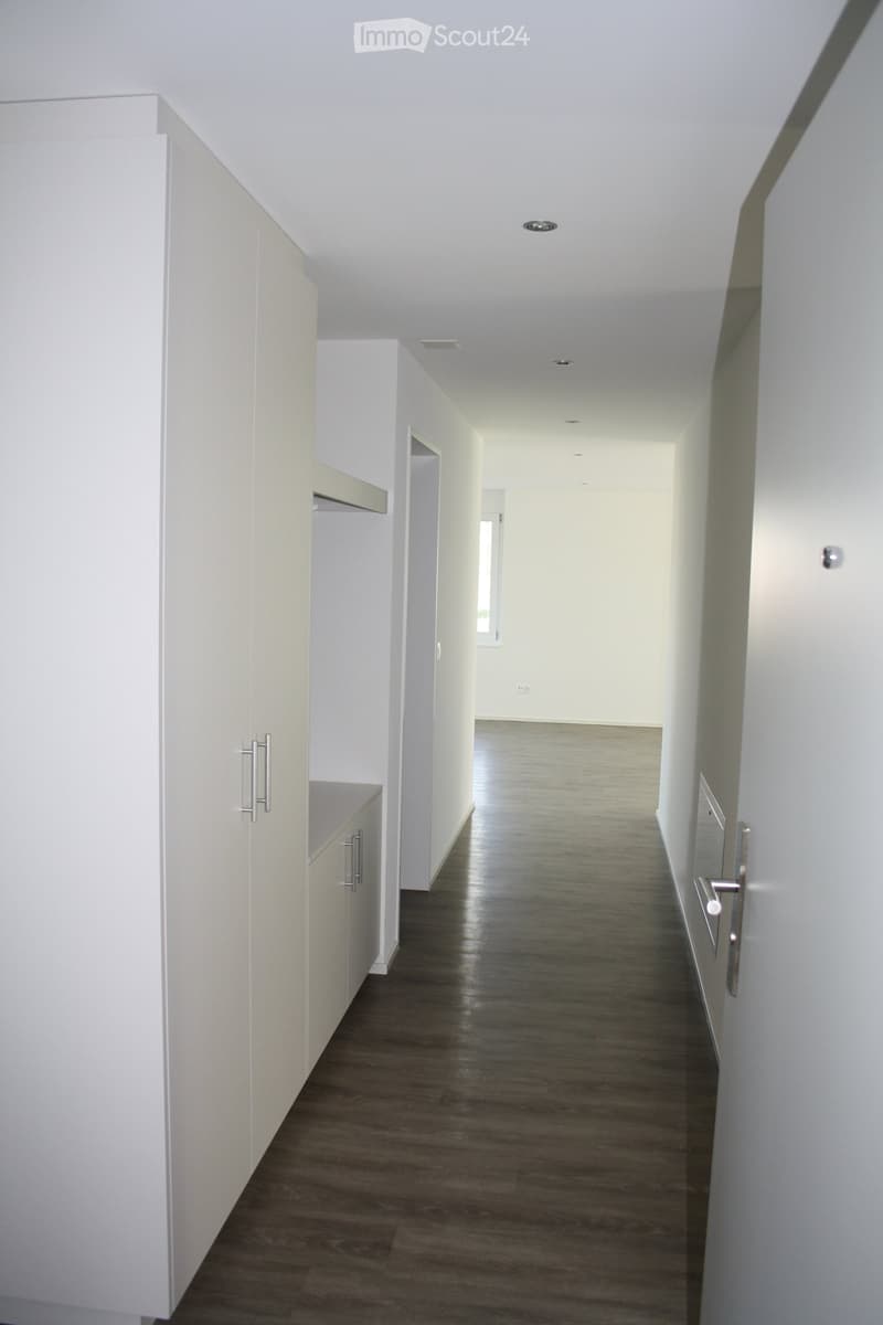Empty hallway with light-colored walls, dark wooden flooring, and white cabinetry on one side.
