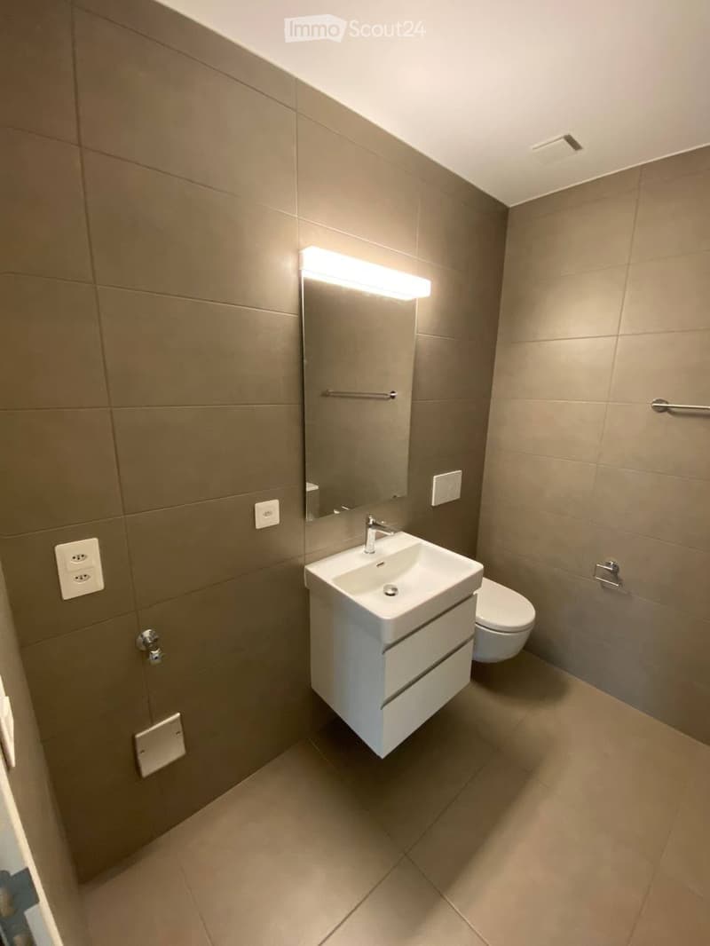 Modern bathroom with beige tiles, white sink, mirror with built-in lighting, and toilet.