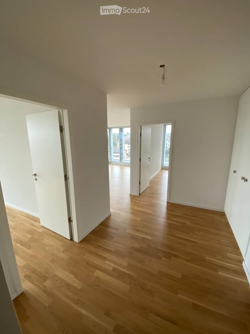 Empty room with light wooden flooring, white walls, and a single ceiling light fixture. The room has two open doors leading to other spaces.