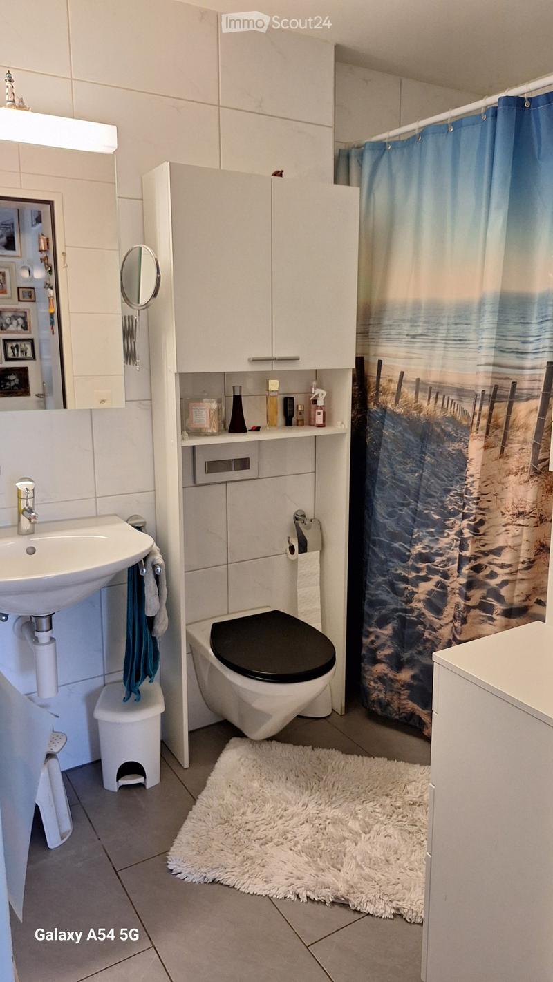 Modern bathroom with white tiles, a sink, toilet, mirror, cabinet, and a beach-themed shower curtain. A white fluffy rug is on the floor, and a small stool is near the sink.