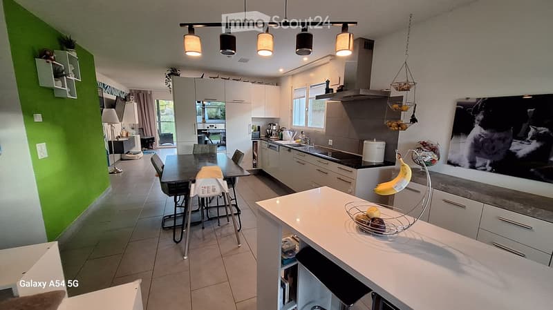 Modern kitchen with white cabinets, a central island, and a dining area. Features include a black countertop, hanging pendant lights, and a large window with a view of the outdoor area.