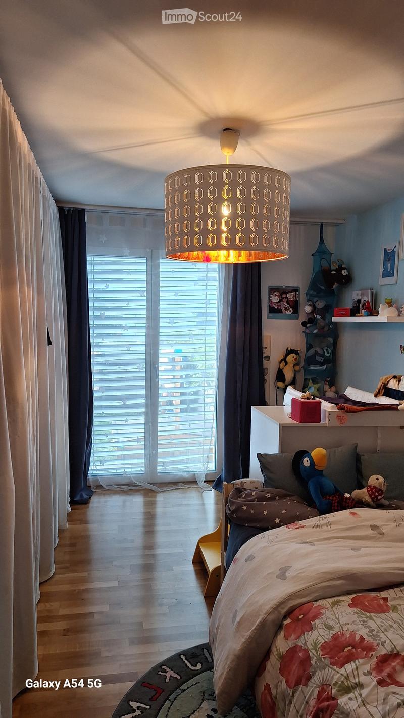 Brightly lit bedroom with wooden flooring, large window with blinds, bed with floral bedspread, decorative pillows, stuffed toys, and a hanging lamp with a geometric shade.
