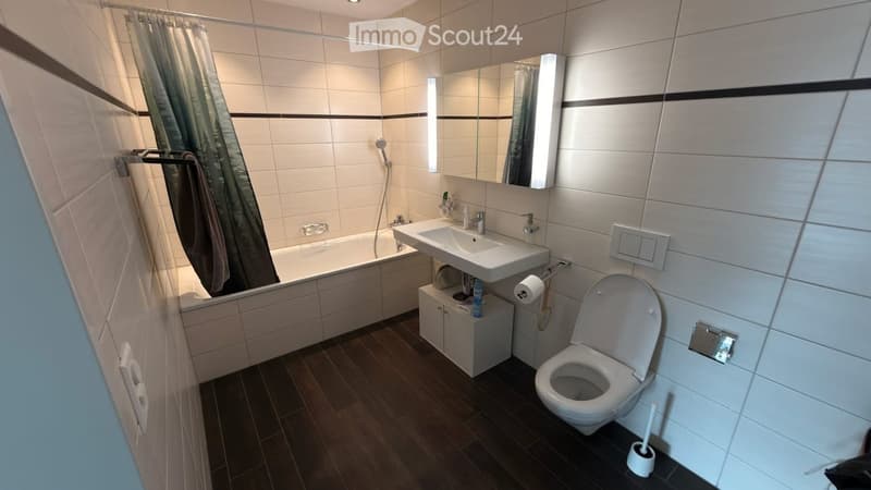 Modern bathroom with a bathtub, sink, toilet, mirror, and a shower curtain. The floor is dark wood, and the walls are tiled with white tiles and black accents.